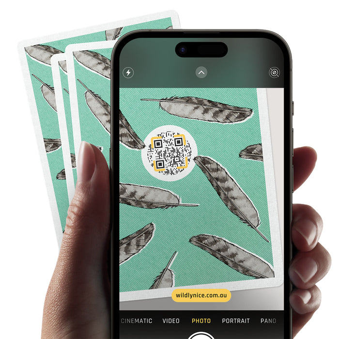 A hand holds a smartphone showing a camera interface with a feather pattern and a QR code, with two matching patterned cards fanned behind it on a white background.