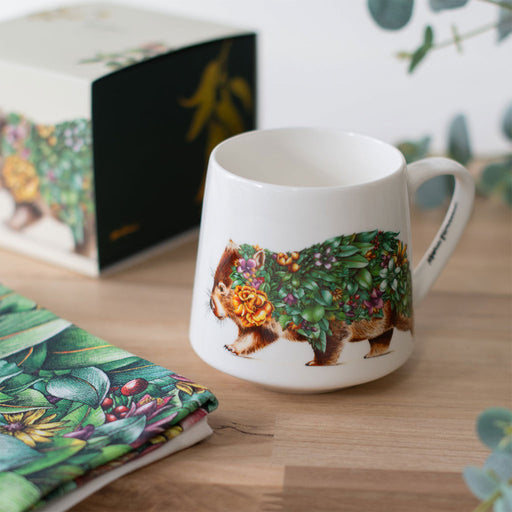 White mug with a decorative wombat design on a wooden surface, with a floral notebook in the background.