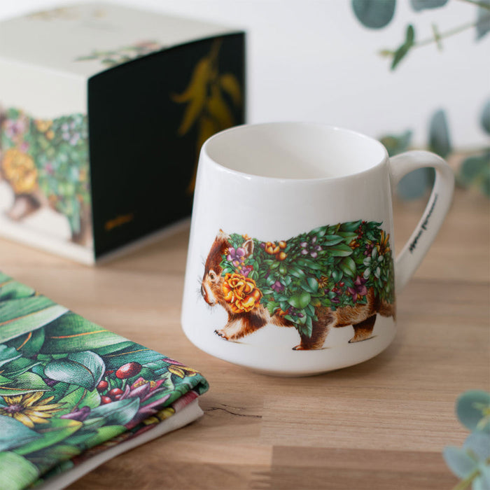 White mug with a decorative wombat design on a wooden surface, with a floral notebook in the background.