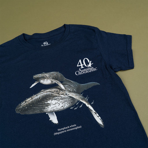 A navy blue short-sleeve T-shirt laid flat on a solid olive-green background. The shirt features a white graphic print of two humpback whales, along with the text '40 Australian Geographic' and 'Humpback whale (Megaptera novaeangliae)'.